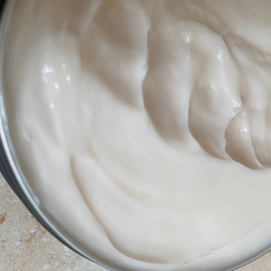 Close-up of a tin filled with creamy white moisturiser on a textured surface.